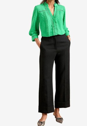 Woman wearing green textured button-up blouse, black wide-leg trousers with cuffed hems, and leopard print pointed flats, hands in pockets.