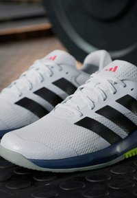 Zapatillas deportivas blancas con tres rayas negras, textura de malla y acentos rojos en la lengüeta. Presentan una suela gris y azul.