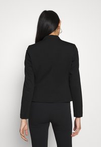 Blazer noir ajusté avec manches longues et col cranté, fabriqué en tissu lisse. Présente une couture au dos et une silhouette cintrée.