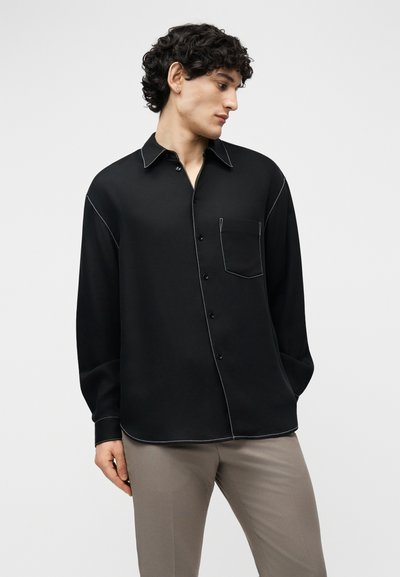 Homme aux cheveux bouclés portant une chemise noire à boutons avec des coutures blanches et un pantalon beige, regardant vers sa gauche sur un fond uni.