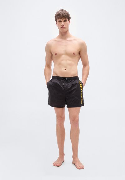 Μαύρο μαγιό σόρτς με οριζόντιες ρίγες, ελαστική μέση, κλείσιμο με κορδόνι και κίτρινο λογότυπο τυπωμένο στο πλάι.