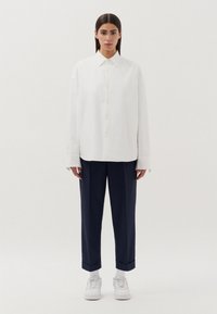 Camicia bianca con colletto strutturato, abbinata a pantaloni scuri blu, cropped e svasati, e sneakers bianche. Design semplice e pulito.