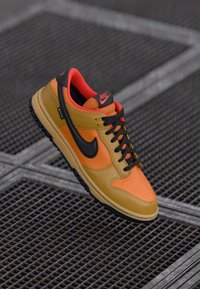 Orange and tan Nike sneaker with black swoosh and laces, red inner lining, resting on metal grid surface.