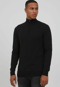 Schwarzer Zip-Kragen-Pullover mit langen Ärmeln und gerippten Bündchen, aus einem weichen Strickmaterial gefertigt. Er weist ein dezentes Nahtdetail auf und hat eine figurbetonte Passform.