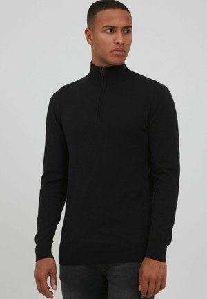 Suéter negro con cuello de cremallera y mangas largas, hecho de un material de punto suave, presenta un sutil detalle de costura y un diseño ajustado.
