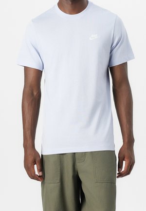 Mężczyzna ubrany w gładką białą koszulkę Nike z krótkim rękawem i oliwkowozielone spodnie, stoi z rękami swobodnie opuszczonymi wzdłuż ciała.
