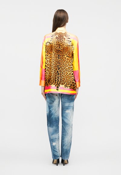 Γυναίκα που φοράει πολύχρωμη μπλούζα με μακριά μανίκια και μεγάλο τύπωμα λεοπάρ στην πλάτη, συνδυασμένη με ανοιχτόχρωμα τζιν με φθορές και μαύρες γόβες.