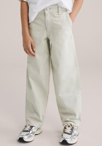 Barn iført løstsiddende, lyse beige bukser med forlommer og hvide sneakers med sølv- og sorte accenter.