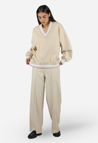 Bež oversize sweatshirt z v-izrezom in belimi detajli, v kombinaciji z bež širokimi hlačami. Oboji oblačili imata mehak teksturo.