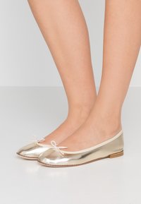 Bailarinas metálicas doradas con punta redondeada, ribete color crema y un pequeño detalle de lazo en la parte delantera. Suela plana y textura suave.