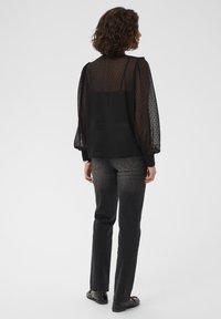 Woman with curly hair wearing sheer black blouse with polka dots, dark gray jeans, and black loafers, standing facing away on white background.
