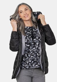 Schwarze Pufferjacke mit glänzender Oberfläche, ausgestattet mit Kapuze, Reißverschluss und grauem Futter. Darunter ein schwarzes Oberteil mit einem weißen Muster.