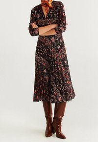 Robe florale noire à manches longues, col en V, et jupe évasée, ornée de motifs paisley rouges et crème. Portée avec des bottes marron montant jusqu'aux genoux.