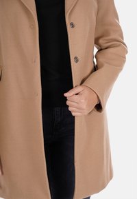 Cappotto di lana color sabbia con un design aderente, dotato di colletto, due bottoni e apertura frontale. Indossato sopra un maglione nero e denim.