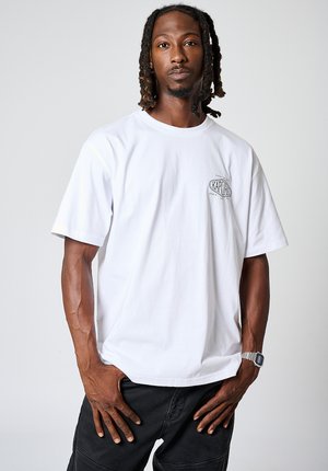 Witte katoenen T-shirt met korte mouwen en een ronde hals. Heeft een kleine zwarte grafische afbeelding op de linkerborst. Draagt een zwarte broek.