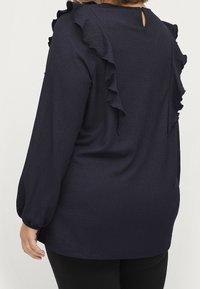 Granatowa bluzka z długim rękawem o fakturowanej tkaninie, z falbankami na ramionach i wycięciem w kształcie klucza na plecach.