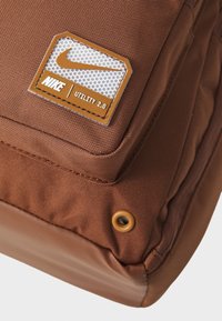 Brązowa torba użytkowa z tkaniny o teksturowanej powierzchni; posiada naszywkę z logo Nike oraz detal w postaci oczka na dole.