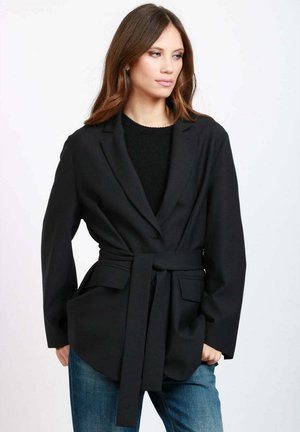 Blazer nero con cintura, dal taglio sartoriale, con colletto a revers, due tasche frontali e maniche lunghe. Indossato sopra un maglione scuro e jeans.