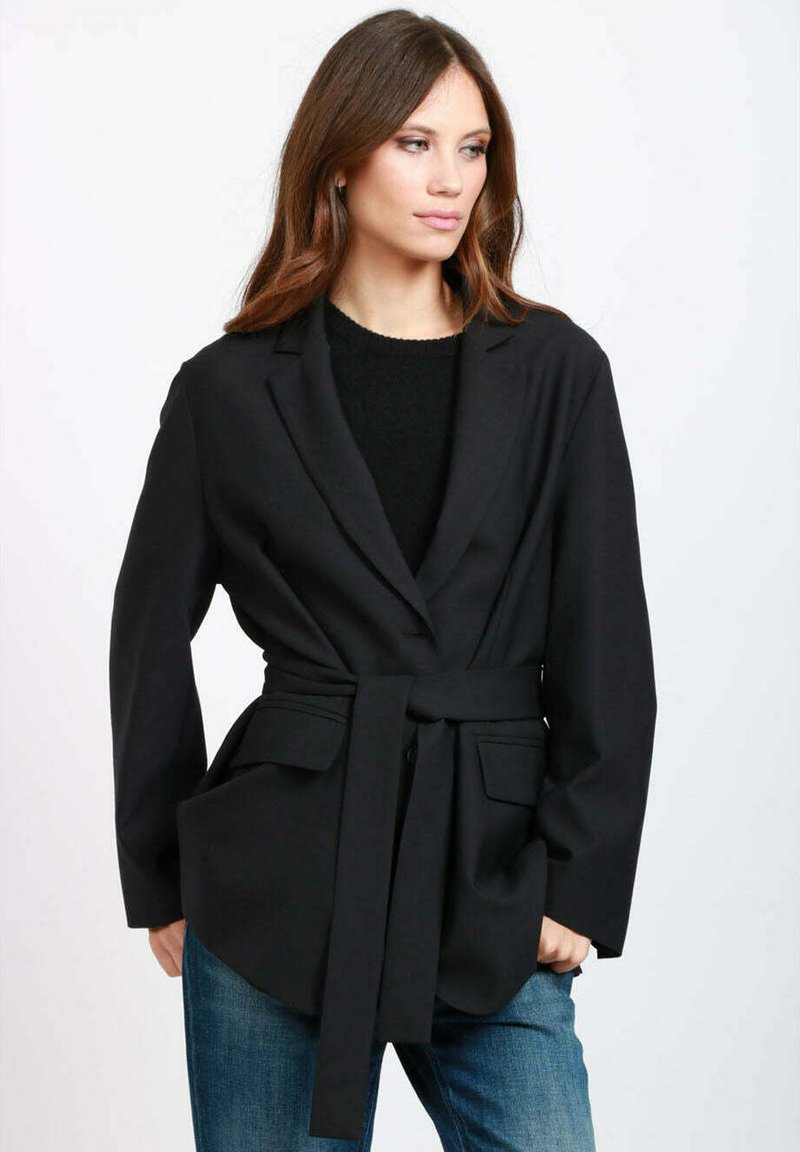 Blazer nero con cintura, dal taglio sartoriale, con colletto a revers, due tasche frontali e maniche lunghe. Indossato sopra un maglione scuro e jeans.