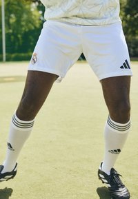 Hvide fodboldshorts med Real Madrid-logo, parret med sort og hvidstribede strømper og sorte fodboldstøvler med struktureret overflade.