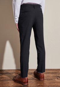 Czarne, dopasowane spodnie o gładkiej fakturze, z dwoma tylnymi kieszeniami zapinanymi na guziki, zestawione z brązowymi butami typu wingtip na drewnianej podłodze.