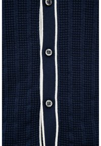 Gebreid marineblauw vest met een witte gestreepte patte, een gestructureerd patroon en vijf donkere knopen die centraal zijn uitgelijnd.