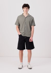 Camisa de botón de manga corta en verde oliva con tejido texturizado, combinada con pantalones cortos de mezclilla negros y zapatillas blancas. Sin patrones ni acentos visibles.