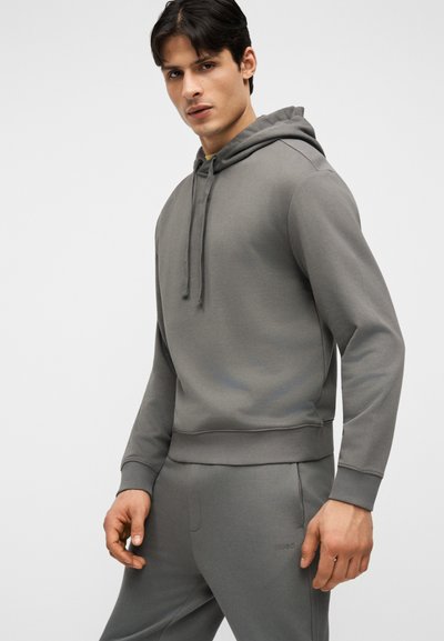 Homme debout, portant un sweat à capuche gris assorti et un pantalon de jogging, regardant légèrement vers la gauche sur un fond clair uni.