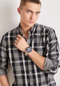 Chemise à carreaux noir et blanc avec col boutonné, accents gris sur les manches, portée avec une montre bleue à bracelet métallique et un cadran détaillé.
