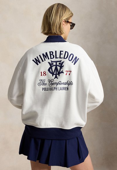 Person with short blonde hair wearing sunglasses, a white jacket with "Wimbledon 1877" and "Polo Ralph Lauren" text, and a navy pleated skirt.