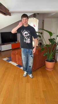 Fekete grafikai mintás póló koponyás dizájnnal, szakadt világos mosott farmer foltokkal, és ezüstszínű sportcipők. Beltéri környezet növényekkel és bútorokkal.