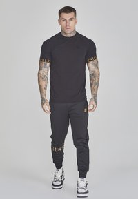 Zwart T-shirt met korte mouwen met goudkleurige branding op de manchetten, gecombineerd met zwarte joggingsbroeken met een zijlogo en gouden tekst, afgewerkt met zwarte sneakers.