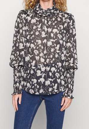 Kvinde iført en gennemsigtig sort bluse med hvide blomsterprint, høj hals, puffede ærmer og blå jeans.