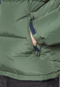 Doudoune verte avec une texture matelassée, poches latérales zippées et poignets côtelés pour un ajustement confortable. Le tissu principal semble résistant à l'eau.