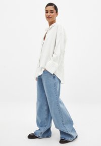 Camicia bianca a righe con bottoni e maniche lunghe, abbinata a jeans a gamba larga di colore blu chiaro e scarpe nere. Design semplice, vestibilità comoda.