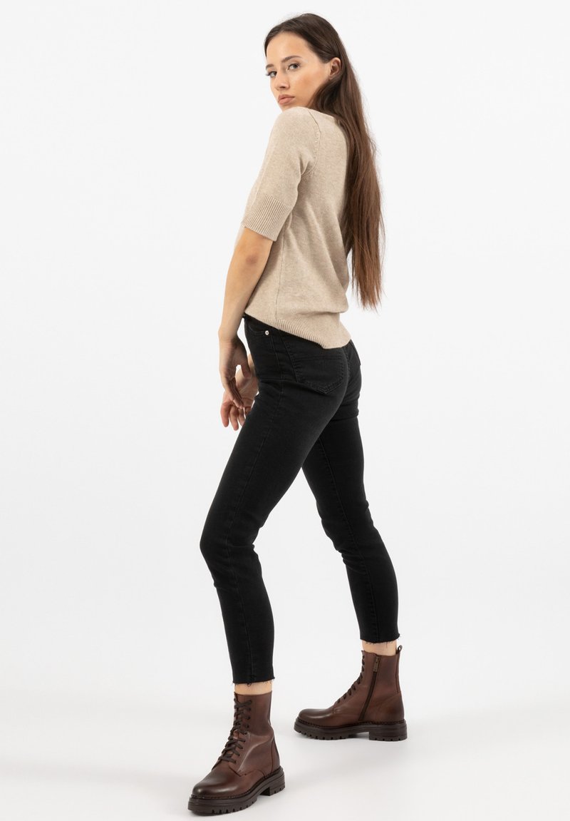 Maglione beige a maniche corte, design attillato, orlo a costine; abbinato a jeans skinny neri e stivaletti alla caviglia marroni con lacci. Sfondo bianco uniforme.