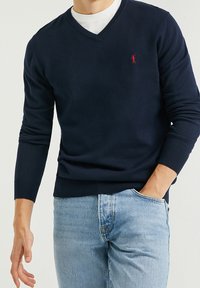Pull col en V bleu marine en tissu maille douce, avec poignets et ourlet côtelés, orné d'un petit logo rouge sur le côté gauche. Porté sur une chemise blanche.