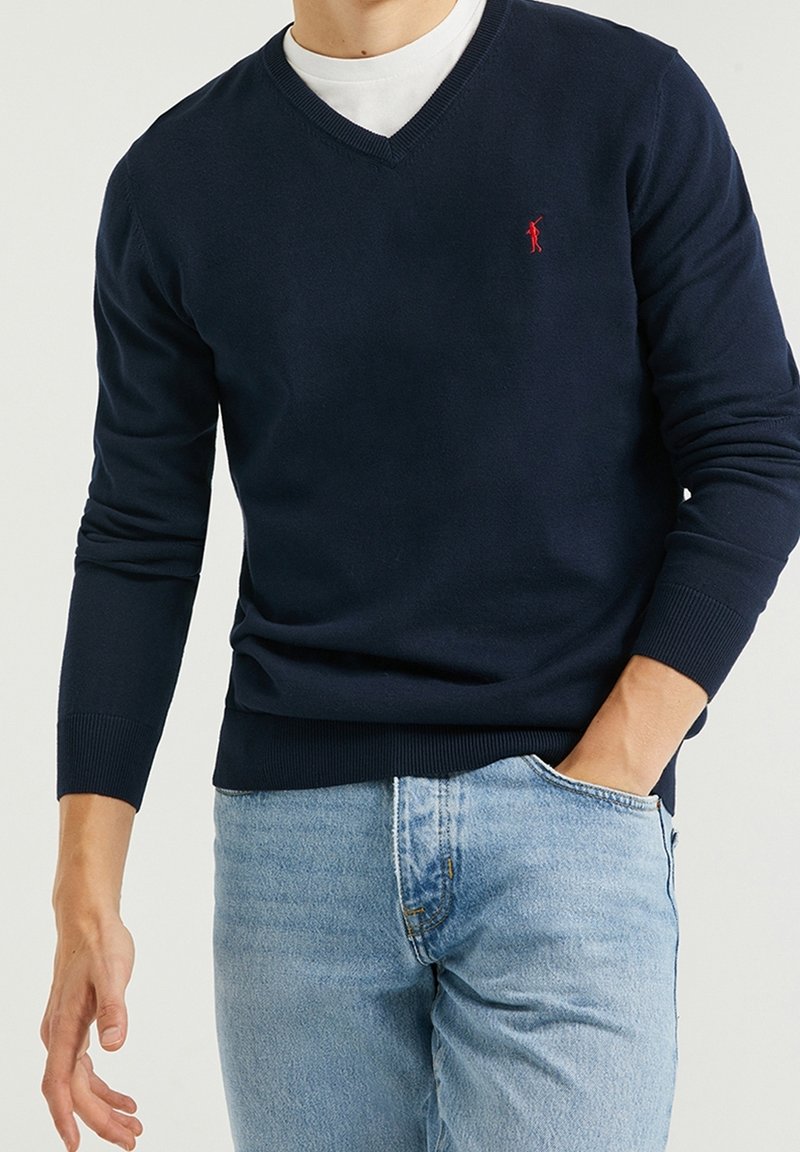 Pull col en V bleu marine en tissu maille douce, avec poignets et ourlet côtelés, orné d'un petit logo rouge sur le côté gauche. Porté sur une chemise blanche.