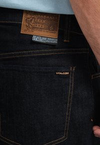 Los jeans de mezclilla azul oscuro tienen un parche de cuero en la cintura con un gráfico y un logotipo, costuras amarillas y una etiqueta de ahorro de agua.