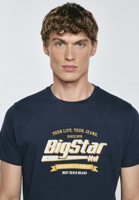 Tamsiai mėlyna medvilninė marškinėliai su baltu ir geltonu spausdintu logotipu bei tekstu. Apvalus kaklas, standartinis sukirpimas, matomi siūlių detalės.