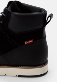 Zwarte leren en suede enkellaars met grijze veters, een gevoerde schacht en een rood Levi's logo nabij de hak, met een gestructureerde witte zool.