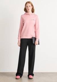 Sudadera de algodón rosa con un bolsillo delantero y logo, combinada con pantalones de pierna ancha negros y zapatillas rosas. Diseño simple y casual.