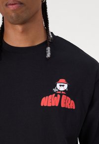 Μαύρο μπλουζάκι με μακριά μανίκια και έναν καρτούν χαρακτήρα του μπέιζμπολ, καθώς και με το λογ ότυπο "NEW ERA" εκτυπωμένο σε κόκκινο στην αριστερή πλευρά του στήθους. Μαλακό βαμβακερό υλικό.