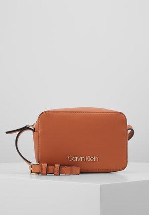 Sac bandoulière rectangulaire en cuir camel Calvin Klein avec logo doré et bandoulière réglable sur une surface blanche.
