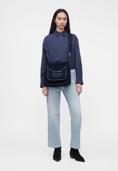 Woman wearing a navy button-up shirt and light blue jeans, carrying a large black shoulder bag with a wide strap, standing against a plain background.