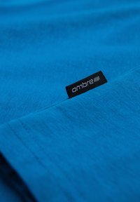 Tessuto di cotone blu con una trama liscia, caratterizzato da un'etichetta nera con il marchio "ombre". Il materiale è piegato, mostrando bordi netti.