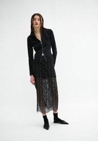 Black ribbed zip-up jacket paired with a semi-transparent black lace skirt featuring circular patterns. Model wears black socks and pointed flats.
