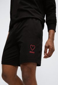 Μαύρο βαμβακερό σορτς με ελαστική μέση. Διαθέτει κόκκινο γραφικό καρδιάς και κείμενο "HUGO" στο κάτω αριστερό μέρος.