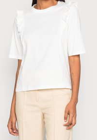 Bílý bavlněný tričko s volánovými detaily na ramenou, krátkými rukávy a volným střihem, spárované s béžovými elegantními kalhotami.