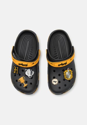 Black clogs with an orange strap feature Hufflepuff and animal-themed accessories. Holes for ventilation and textured footbed provide comfort.